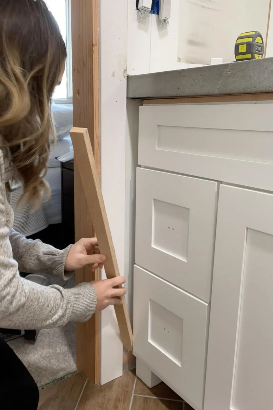 Woman holding a wooden trim piece next to a white shaker-style bathroom cabinet to measure and align it for installation during renovation.